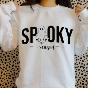 K&ouml;nnte beinhalten: Wei&szlig;es Sweatshirt mit den W&ouml;rtern "SPOOKY" und "season" in Schwarz. Eine Geistergrafik befindet sich in der Mitte des Wortes "SPOOKY". Das Sweatshirt ist ein Halloween-Themenartikel.