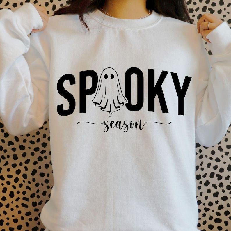 K&ouml;nnte beinhalten: Wei&szlig;es Sweatshirt mit den W&ouml;rtern "SPOOKY" und "season" in Schwarz. Eine Geistergrafik befindet sich in der Mitte des Wortes "SPOOKY". Das Sweatshirt ist ein Halloween-Themenartikel.