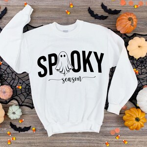K&ouml;nnte beinhalten: Wei&szlig;es Sweatshirt mit dem Schriftzug "Spooky Season" in Schwarz und einem Geistermotiv. Das Sweatshirt ist von Halloween-Dekorationen umgeben, darunter K&uuml;rbisse, Flederm&auml;use und Spinnweben. Ein festlicher und saisonaler Bekleidungsartikel.