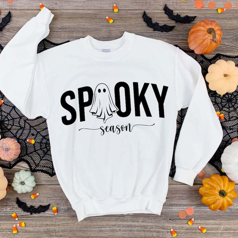 K&ouml;nnte beinhalten: Wei&szlig;es Sweatshirt mit dem Schriftzug "Spooky Season" in Schwarz und einem Geistermotiv. Das Sweatshirt ist von Halloween-Dekorationen umgeben, darunter K&uuml;rbisse, Flederm&auml;use und Spinnweben. Ein festlicher und saisonaler Bekleidungsartikel.