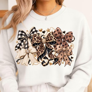 May include: A white long-sleeve sweatshirt featuring a central graphic print of multiple decorative bows. The bows include patterns of black and white buffalo plaid, leopard print, and solid tan with black hearts and stars, accented by gold charms.