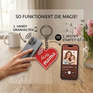 Könnte beinhalten: Ein roter herzförmiger Schlüsselanhänger mit den weißen Worten "beste Mama", befestigt an einem silbernen Schlüsselring. Ein Smartphone wird in der Nähe des Schlüsselanhängers gehalten, mit dem Text "SO FUNKTIONIERT DIE MAGIE!" darüber. Ein zweites Telefon zeigt eine Spotify-Playlist.