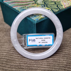 Könnte beinhalten: Ein transluzentes, hellgrünes Jadeit-Armreif-Armband. Das Armband ist kreisförmig und glatt, mit polierter Oberfläche. Ein Etikett mit dem Text "NATURAL JADEITE JADE" und "P-lab" ist angebracht. Der Hintergrund zeigt eine dekorative Box.