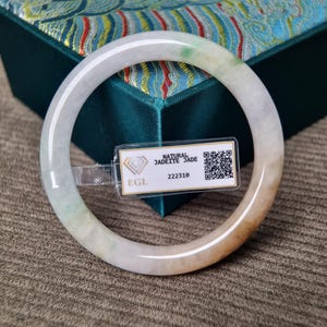 May include: A smooth, circular jadeite bangle featuring translucent white, pale green, and warm amber tones. A rectangular tag is attached to the bangle, displaying the text "NATURAL JADEITE JADE", "EGL", the number "222310", and a QR code.