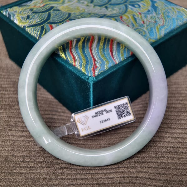 May include: A smooth, round jadeite bangle featuring pale green and soft lavender hues. A small white rectangular tag is attached to the bangle, displaying the text NATURAL JADEITE JADE, the number 222643, the letters EGL, and a QR code.