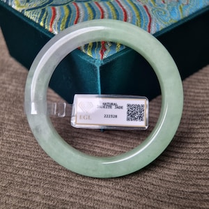 May include: A smooth, pale green jadeite bangle bracelet with a translucent appearance. A small white rectangular tag is attached to the bangle, featuring the text NATURAL JADEITE JADE, EGL, 222328, and a QR code printed on the surface.