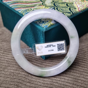 Peut inclure: Un bracelet jonc en jade jadéite translucide. Le bracelet est principalement blanc avec de subtiles nuances de vert et de gris. Le bracelet est présenté devant une boîte turquoise avec un couvercle décoratif. Une étiquette avec du texte est attachée au bracelet.