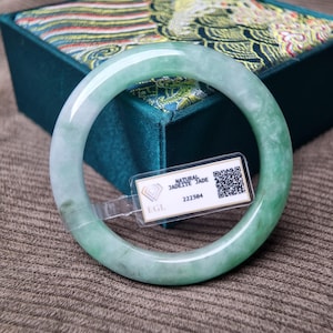 Può includere: Un braccialetto bangle in giada giadeite verde chiaro traslucido. La superficie liscia e lucida del braccialetto presenta un mix di tonalità verde pallido e bianco. Un'etichetta con la scritta "NATURAL JADEITE JADE" e un codice QR è attaccata. Il braccialetto è appoggiato su una superficie marrone.
