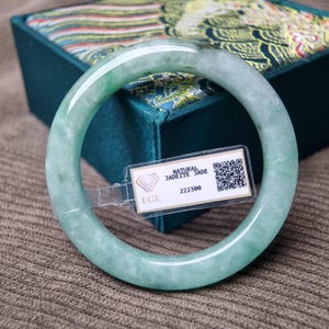 May include: A translucent jadeite jade bangle in shades of green and white. The smooth, circular bracelet is displayed with a tag that reads "NATURAL JADEITE JADE" and the number "222300". The bangle is resting on a textured surface.