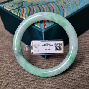 May include: A smooth, circular jadeite bangle with translucent light green and white mottled coloring. A small rectangular white tag is attached to the bangle, featuring the text NATURAL JADEITE JADE, EGL, 222343, and a QR code.