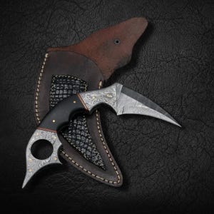 May include: A curved karambit knife with a patterned blade and black handle, resting in a brown leather sheath. The knife features a finger hole and decorative metal accents. The sheath has a textured, dark pattern.