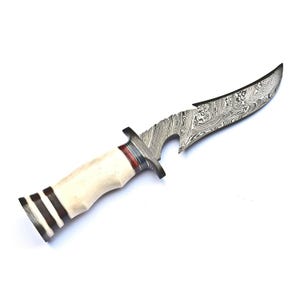 Può includere: Coltello da caccia fatto a mano con una lama curva e decorata. L'impugnatura è bianco sporco con dettagli marrone scuro e rossi. La lama ha un design unico ed è in acciaio.