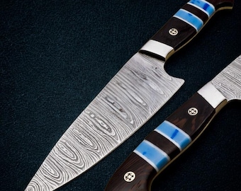 May include: Two chef knives with patterned steel blades and dark wood handles. The handles feature blue and white accents and decorative metal rivets. The blades have a distinctive swirling pattern.