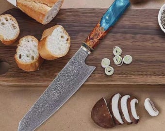May include: A Damascus steel chef's knife with a blue and wood grain handle, resting on a wooden cutting board. Sliced mushrooms, bread, and herbs are arranged around the knife, suggesting culinary use.