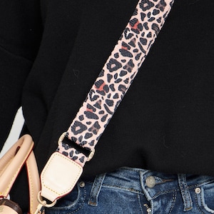 May include: A black sweater with a pink and red leopard print adjustable shoulder strap for a handbag. The strap is attached to a brown leather handbag with a gold clasp. The bag is partially visible and has a brown and tan monogram pattern. The person is wearing blue jeans.