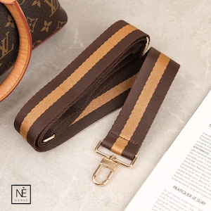 May include: A brown and tan striped bag strap with gold-coloured metal hardware. The strap is made of a durable fabric and features a classic design. The strap is designed to be attached to a handbag or purse.