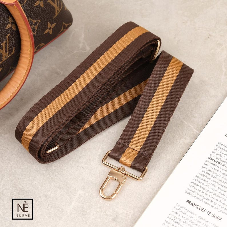May include: A brown and tan striped bag strap with gold-coloured metal hardware. The strap is made of a durable fabric and features a classic design. The strap is designed to be attached to a handbag or purse.