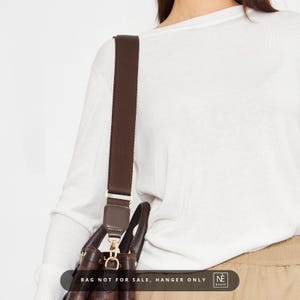 May include: A brown shoulder strap attached to a handbag. The strap is made of a woven material and has gold-coloured metal hardware. The handbag is brown and has a checkered pattern. The text "BAG NOT FOR SALE, HANGER ONLY" is visible.