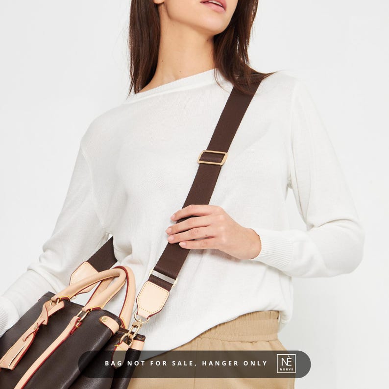 May include: A dark brown handbag with a matching shoulder strap. The strap is adjustable and features gold-coloured metal hardware. The bag has light tan accents and a red interior. The image includes the text: BAG NOT FOR SALE, HANGER ONLY.