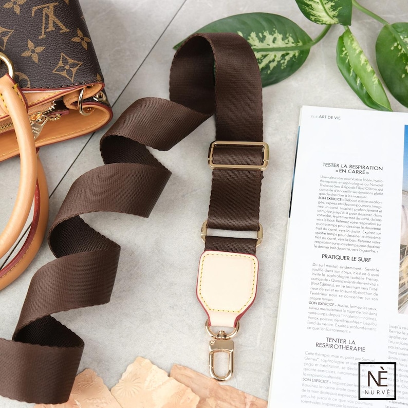 May include: A dark brown nylon bag strap with gold-coloured metal hardware. The strap features a cream-coloured leather accent with a red edge and a gold-coloured clasp. The bag strap is next to a brown and tan handbag.