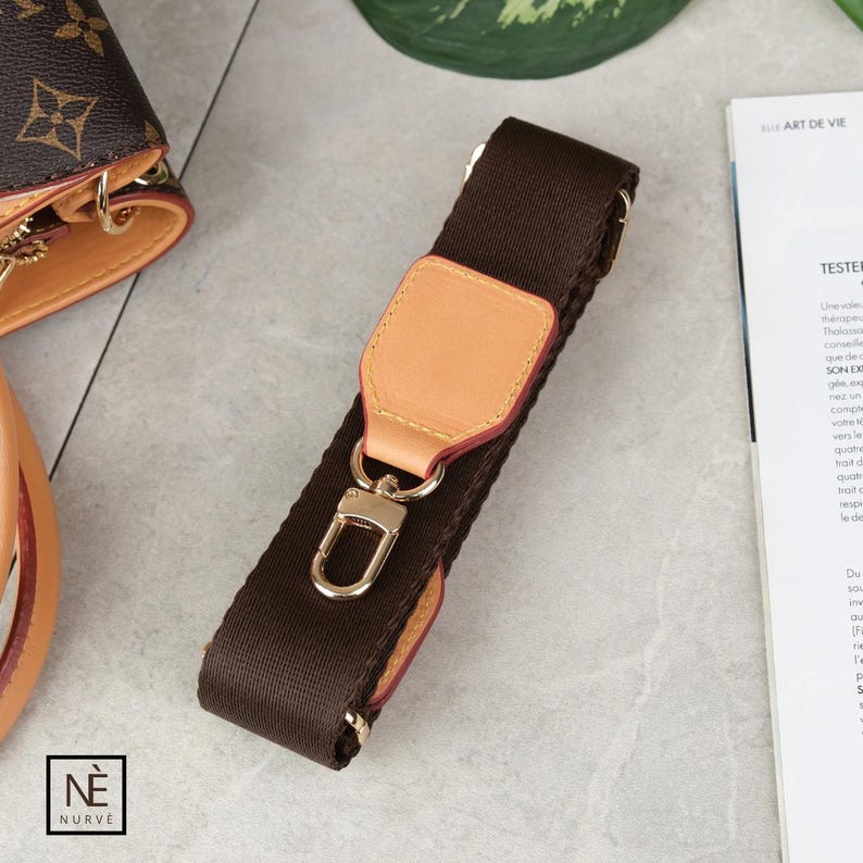 May include: A brown handbag strap with a light tan leather accent and gold-coloured hardware. The strap is made of a woven material and features a metal clasp for attachment. The brand logo is visible in the bottom left corner.