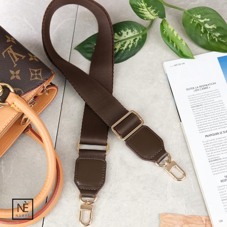 May include: A brown handbag strap with gold-tone metal clasps and buckles. The strap is made of a woven material with leather detailing. The strap is designed to be attached to a handbag. The bag in the image has a brown monogram pattern.