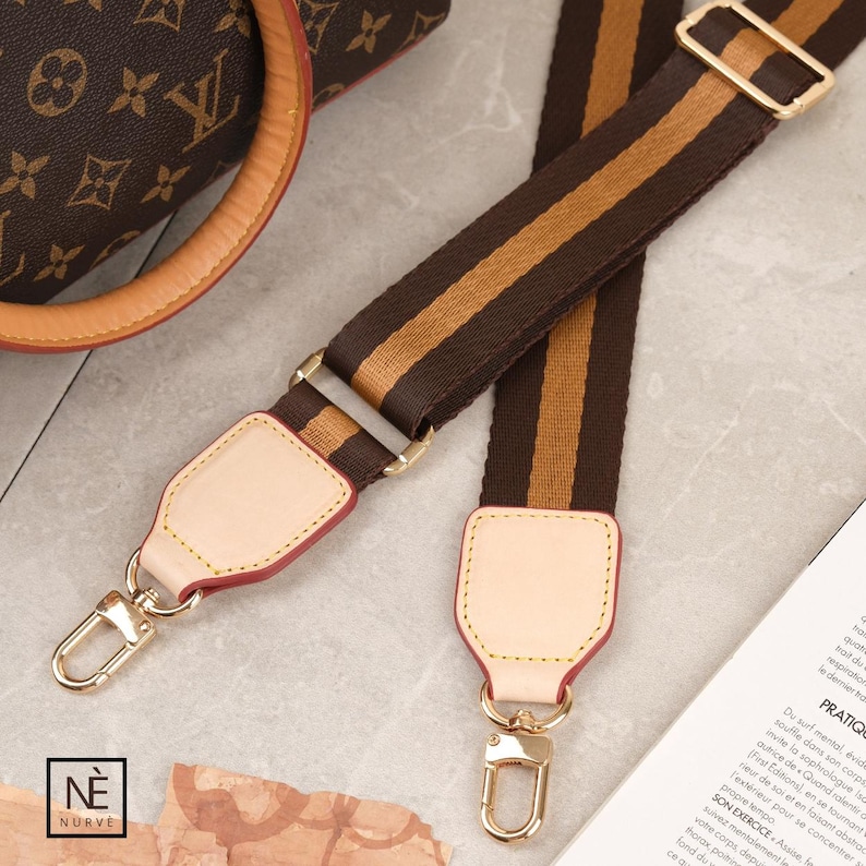 May include: Brown and tan striped handbag strap with gold-tone metal hardware and leather detailing. The strap features a repeating stripe pattern. The strap is attached to a brown handbag.