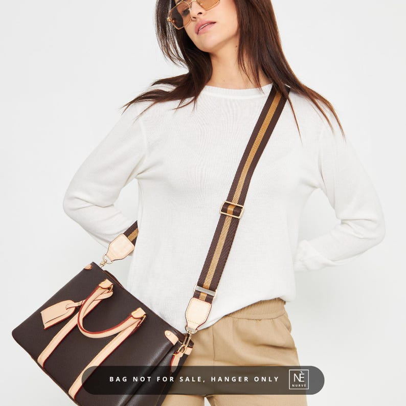May include: A dark brown tote bag with light brown detailing and a striped shoulder strap. The strap has brown and tan stripes. The bag is rectangular with a top handle and a detachable strap. The text "BAG NOT FOR SALE, HANGER ONLY" is visible.