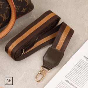 May include: A brown and tan striped bag strap with gold-coloured metal fixings. The strap is made of a woven material and has a leather accent at the end with a gold-coloured clasp. The strap is designed to be attached to a handbag.