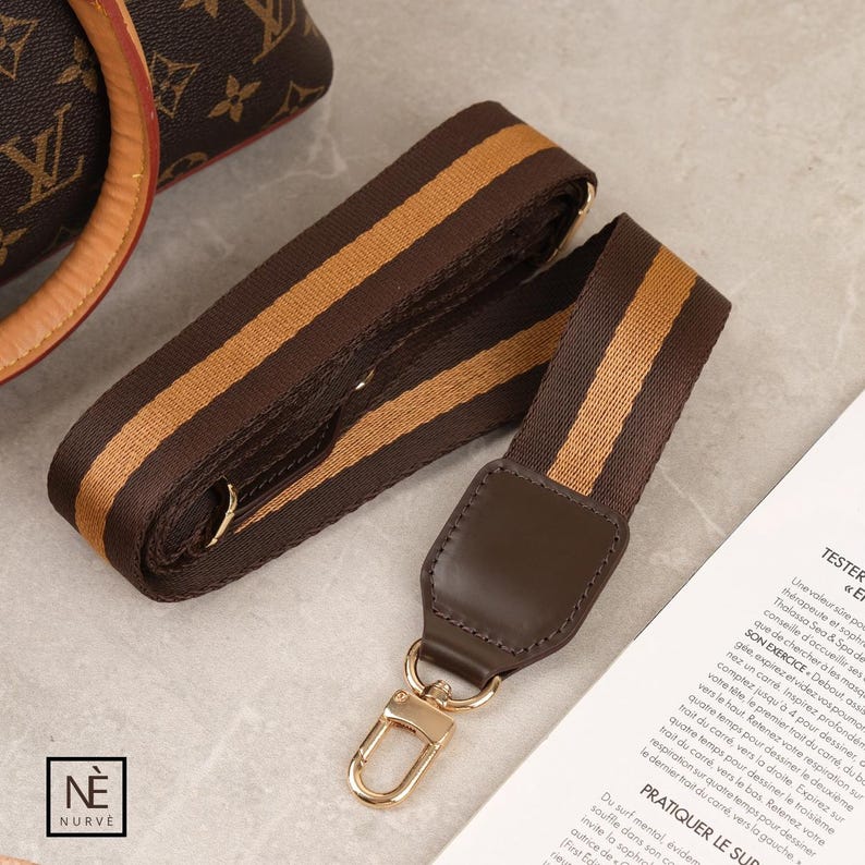 May include: A brown and tan striped bag strap with gold-coloured metal fixings. The strap is made of a woven material and has a leather accent at the end with a gold-coloured clasp. The strap is designed to be attached to a handbag.