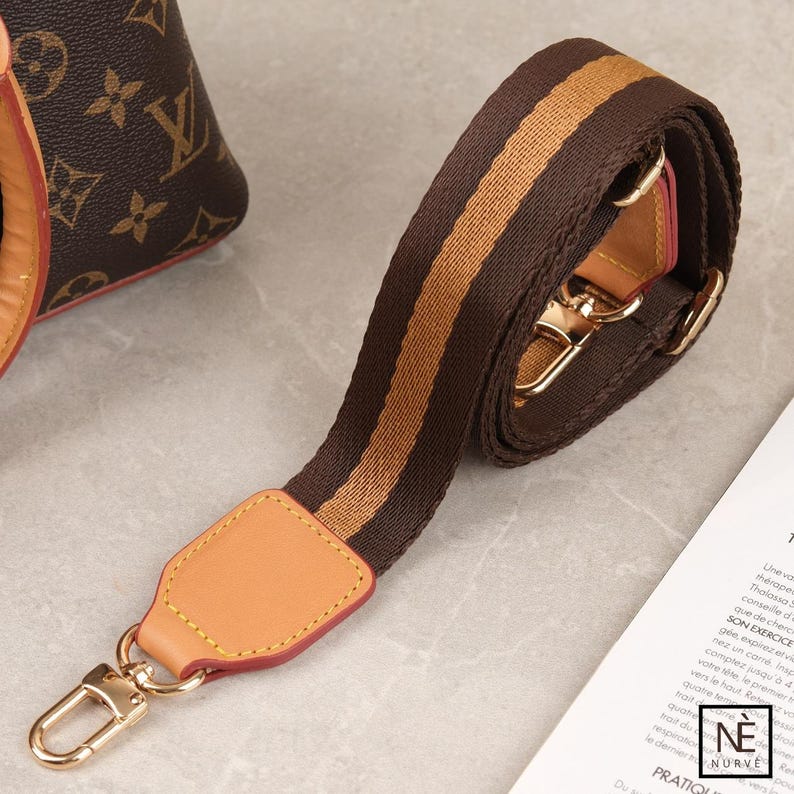 May include: A brown and tan striped bag strap with gold-coloured hardware. The strap has a leather accent with a gold clasp. The strap is designed to be attached to a bag. The background includes a portion of a brown bag.
