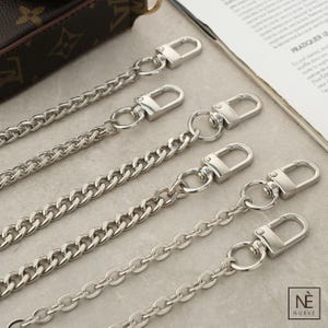 May include: Silver metal purse chains with lobster claw clasps. The chains feature different styles, including curb and oval link designs. The chains are shown on a neutral surface, suitable for accessories and hardware.