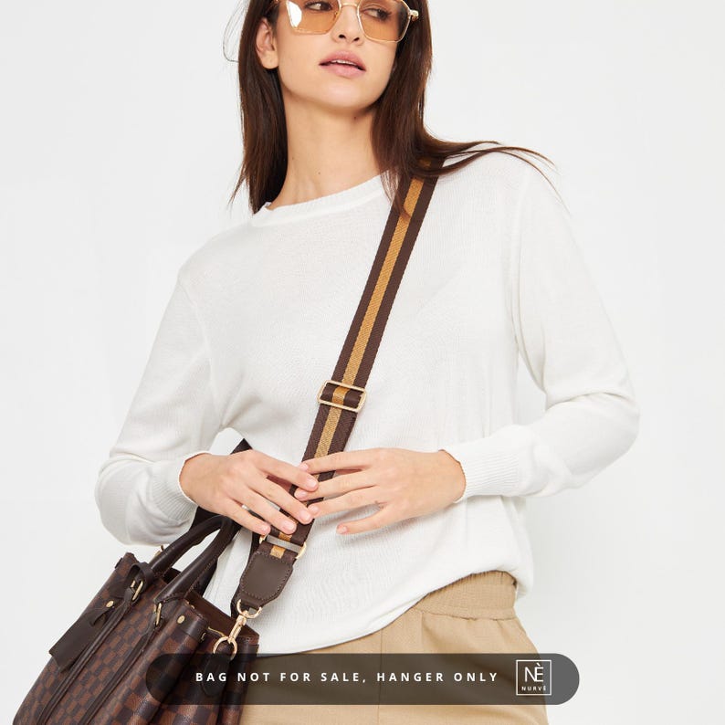 May include: A woman in a white sweater and beige trousers, with a brown and tan striped shoulder strap. The strap is attached to a brown patterned bag. The text "BAG NOT FOR SALE, HANGER ONLY" is visible.