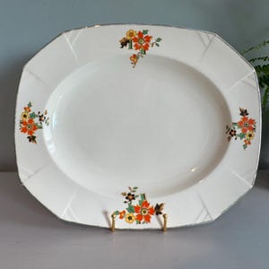 May include: An off-white oval serving platter with a silver rim and a floral design in orange, yellow, and black. The platter has a geometric shape with a slightly raised edge and is displayed on a gold-coloured stand.