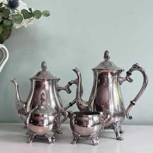 Puede incluir: Un juego de té plateado pulido que incluye una tetera, una jarra de crema, un azucarero y una jarra de servir. El juego presenta detalles elaborados en las asas, las boquillas y los pies. Las superficies reflectantes sugieren un estilo vintage.