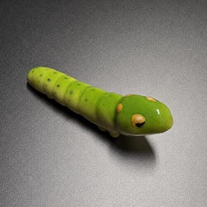 Op de afbeelding: Een klein, glad, limoengroen keramieken rupsfiguurtje met een gesegmenteerd lichaam. Het oppervlak heeft subtiele donkergroene stippen langs de rug en twee opvallende, ronde oranje markeringen op de kop, wat zorgt voor een glanzende afwerking.