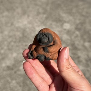 Könnte beinhalten: Eine kleine, handgroße Orang-Utan-Figur mit strukturiertem braunem Fell und einem dunklen, detaillierten Gesicht. Der Primat ist in einer sitzenden, gebeugten Haltung dargestellt, wobei die Arme auf dem Schoß ruhen und feine Schnitzdetails zeigen.