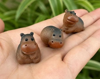 May include: Three miniature brown hippopotamus figurines with smooth, rounded bodies, small ears, and dark, glossy eyes. Each hippo has a slightly darker brown snout and a textured, matte finish, presented as a set of small, hand-sized animal sculptures.