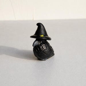 May include: A small black raven figurine wearing a pointed black witch hat. The hat features a single, small, yellow star detail on the front. The bird has textured black feathers, a dark beak, and a glossy eye, standing in a compact pose.