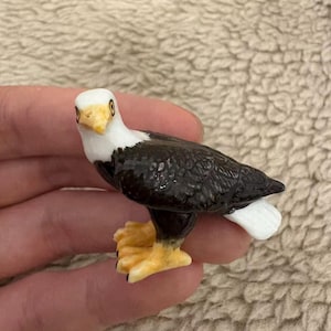May include: A small bald eagle figurine. The eagle has a white head, yellow beak and talons, and black and white feathers. The figurine is detailed and appears to be made of a glossy material.