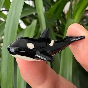 Op de afbeelding: Een kleine, glanzende zwart-witte orka-figuur. De orka heeft witte markeringen en een witte buik. De figuur wordt tussen twee vingers gehouden, met een groene, bladerrijke achtergrond.