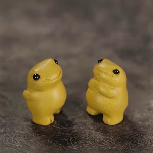 Peut inclure: Deux petites figurines de grenouilles jaune moutarde debout. Chaque grenouille a un corps lisse et arrondi, un sourire subtil et deux petits yeux noirs. Les figurines sont placées côte à côte, montrant leur design anthropomorphe simple.