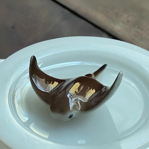 May include: A brown ceramic bird ornament with outstretched wings, placed on a white oval plate. The bird has a glossy finish and a small, dark eye. The plate is smooth and reflective, creating a simple, elegant display.