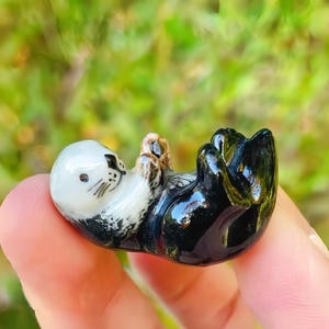 Puede incluir: Figura de cerámica de una nutria marina, tumbada boca arriba. La nutria tiene una cara blanca con bigotes negros y un cuerpo negro. La figura es pequeña y detallada, con un acabado brillante.