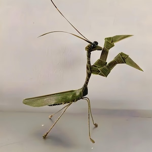 May include: A decorative praying mantis sculpture crafted from woven green palm leaves. The insect figure features long, thin antennae, dark bead eyes, folded front legs, and slender wire-like legs supporting its elongated body structure.