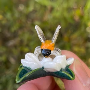 Puede incluir: Una figura de abeja en miniatura hecha a mano, con una flor blanca y hojas verdes. La abeja tiene una cara negra, un pecho naranja y alas plateadas. La flor es blanca con hojas verdes.
