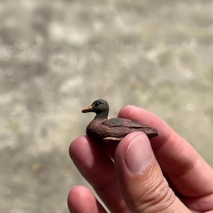 Könnte beinhalten: Eine Miniatur-Entenfigur mit braunem Körper, dunkelgrünem Kopf und orangefarbenem Schnabel. Die kleine Skulptur wird in der Hand einer Person gehalten, wodurch ihre detaillierten Merkmale und ihr realistisches Design hervorgehoben werden. Die Ente ist ein charmantes Dekorationsstück.