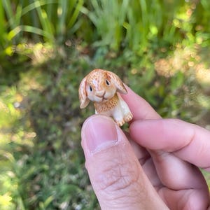 Może przedstawiać: Mała ceramiczna figurka królika z brązowymi i beżowymi detalami futra. Królik ma długie uszy i siedzi. Figurka jest trzymana w dłoni na rozmytym zielonym tle.