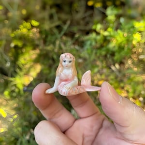 Puede incluir: Pequeña figura de sirena de cerámica, con cola rosa y top melocotón claro. La sirena tiene pelo largo y rubio y está sentada. La figura se sostiene en una mano sobre un fondo verde borroso.