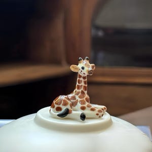 Puede incluir: Una pequeña figura decorativa de una jirafa con manchas marrones y cuerpo color crema. La jirafa está tumbada sobre una base circular blanca. El fondo está borroso, con tonos marrones cálidos.